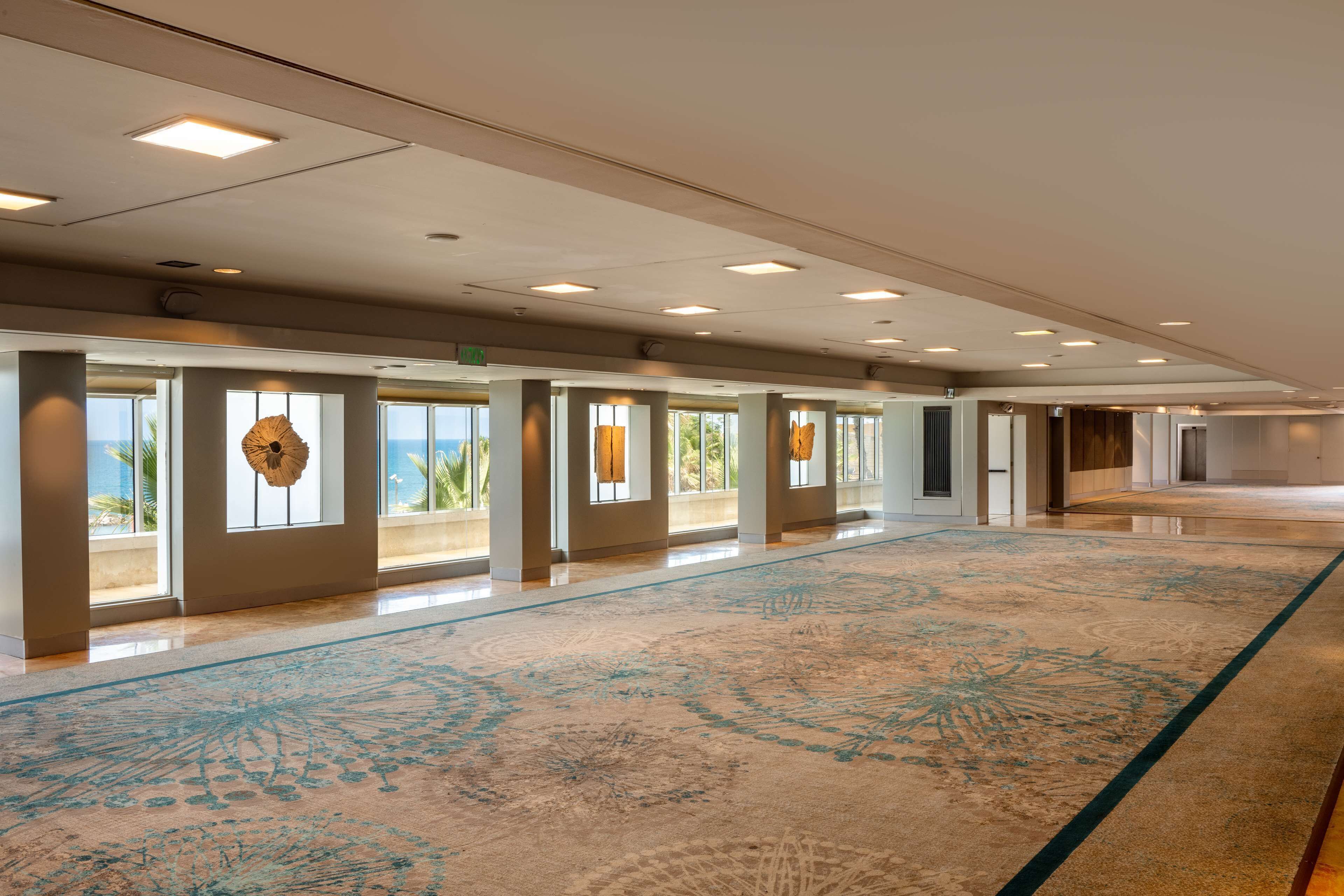 A spacious hotel lobby with a large, patterned carpet, large windows with views, and a modern ceiling design.
