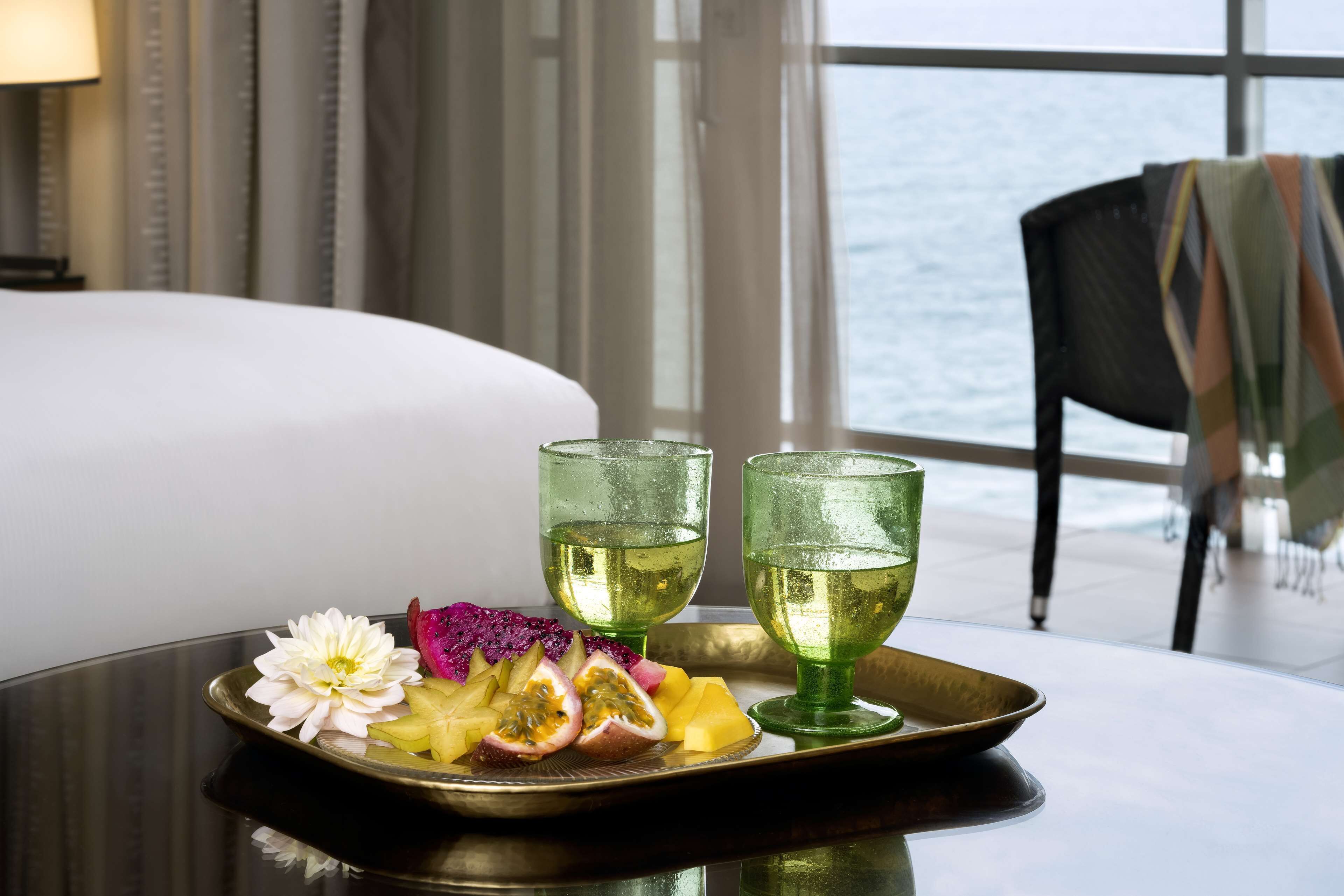 A tray with fruit and two glasses of a clear liquid on a table in a room with a view of the sea.