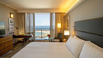 A hotel room with a large bed, a desk with a chair, a television, and a view of the beach.