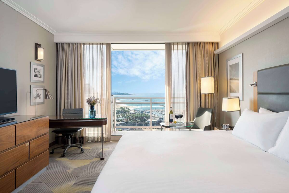 A hotel room with a large bed, a desk, a chair, a TV, a lamp, and a view of the sea.
