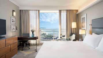 A hotel room with a large bed, a desk, a chair, a TV, a lamp, and a view of the sea.