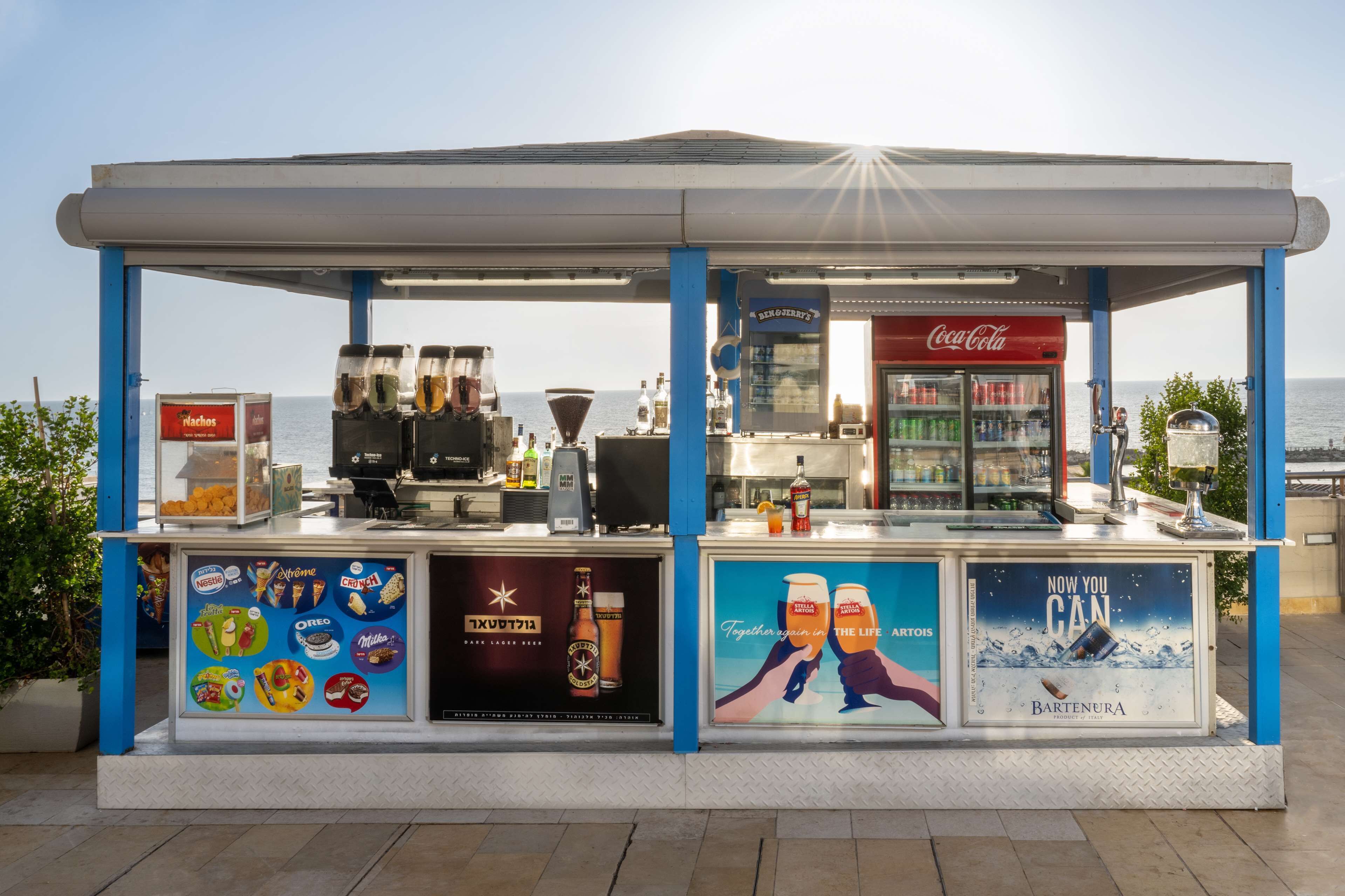 A seaside kiosk with a Coca-Cola refrigerator, a beer tap, and a display of snacks and drinks.