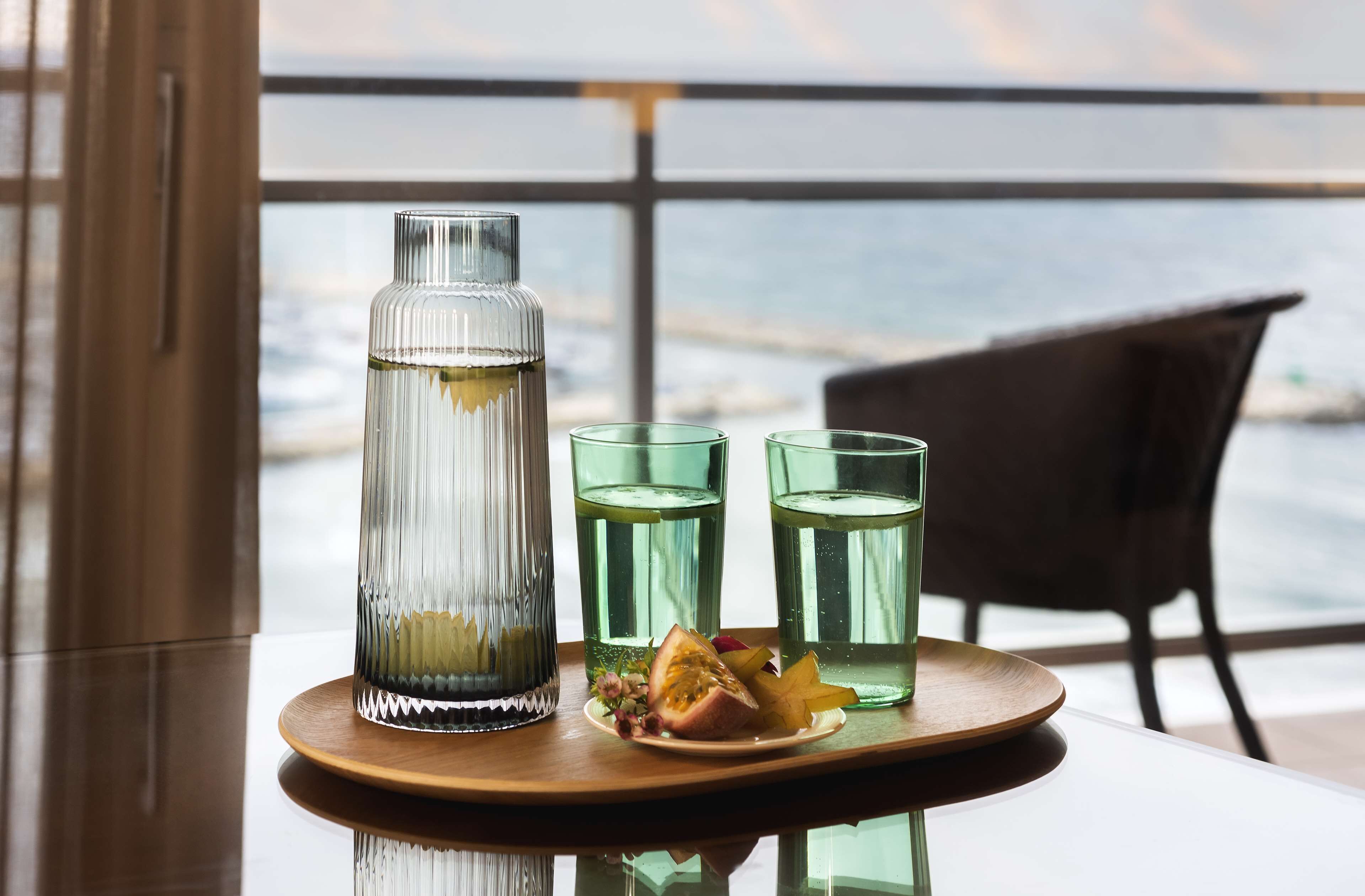A glass bottle and three glasses on a tray with a slice of fruit and flowers, set on a table with a view of the ocean.