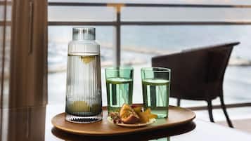 A glass bottle and three glasses on a tray with a slice of fruit and flowers, set on a table with a view of the ocean.