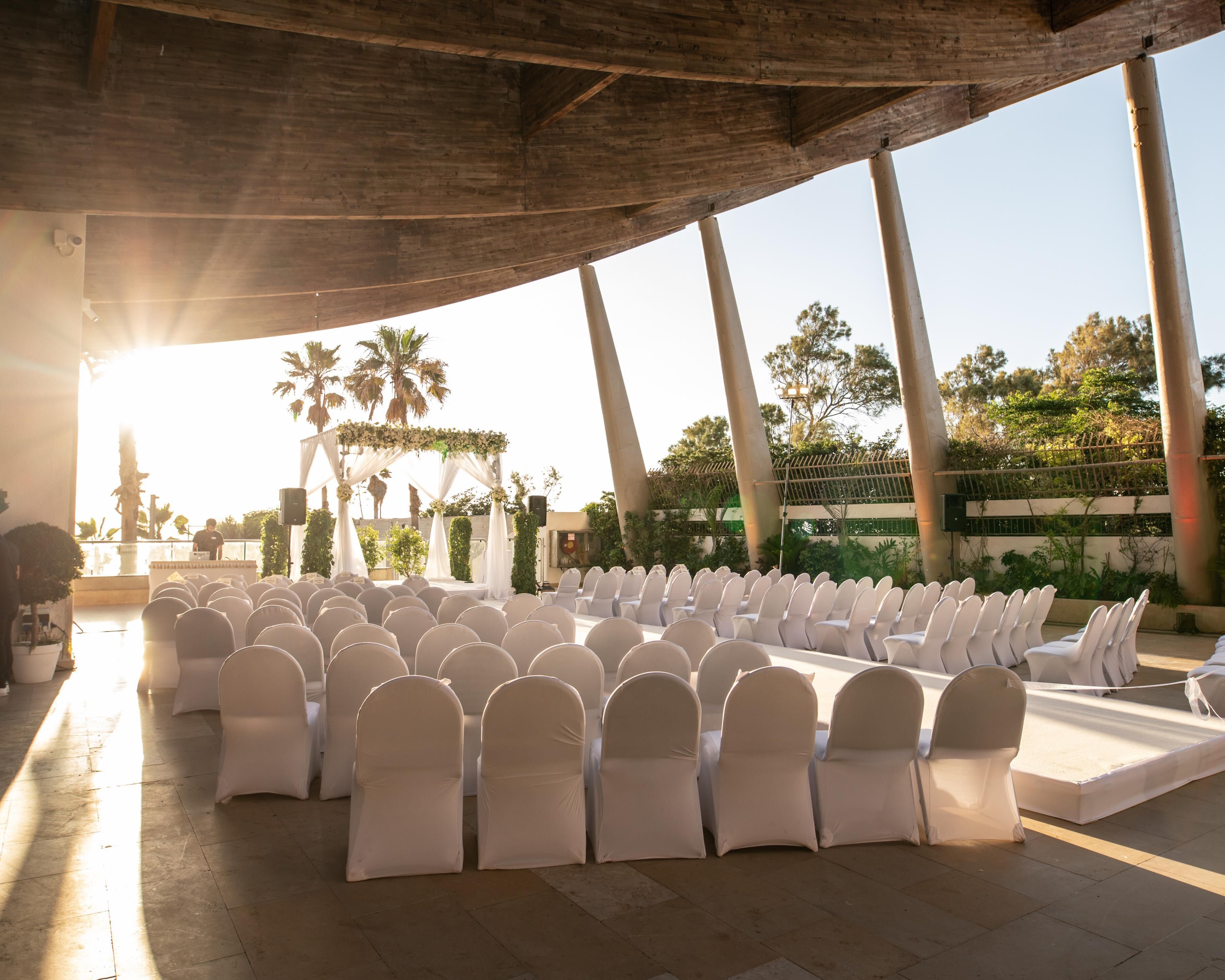 An outdoor event space with rows of white chairs and a stage, surrounded by palm trees and a clear sky.