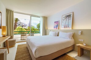 Habitación de hotel con una cama grande, un escritorio, una silla y un balcón con vistas a zonas verdes y edificios.