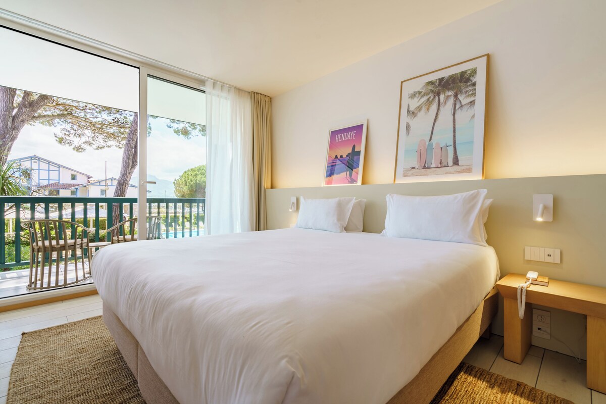 Habitación de hotel con una cama grande, una mesita de noche con un teléfono, una ventana con vistas al exterior y dos cuadros en la pared.