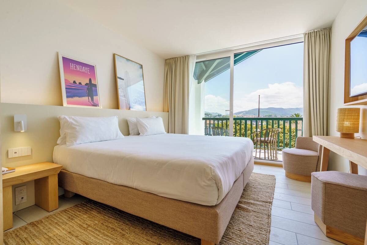 Habitación de hotel con una cama grande, un escritorio, una silla y un balcón con vistas a la ciudad.
