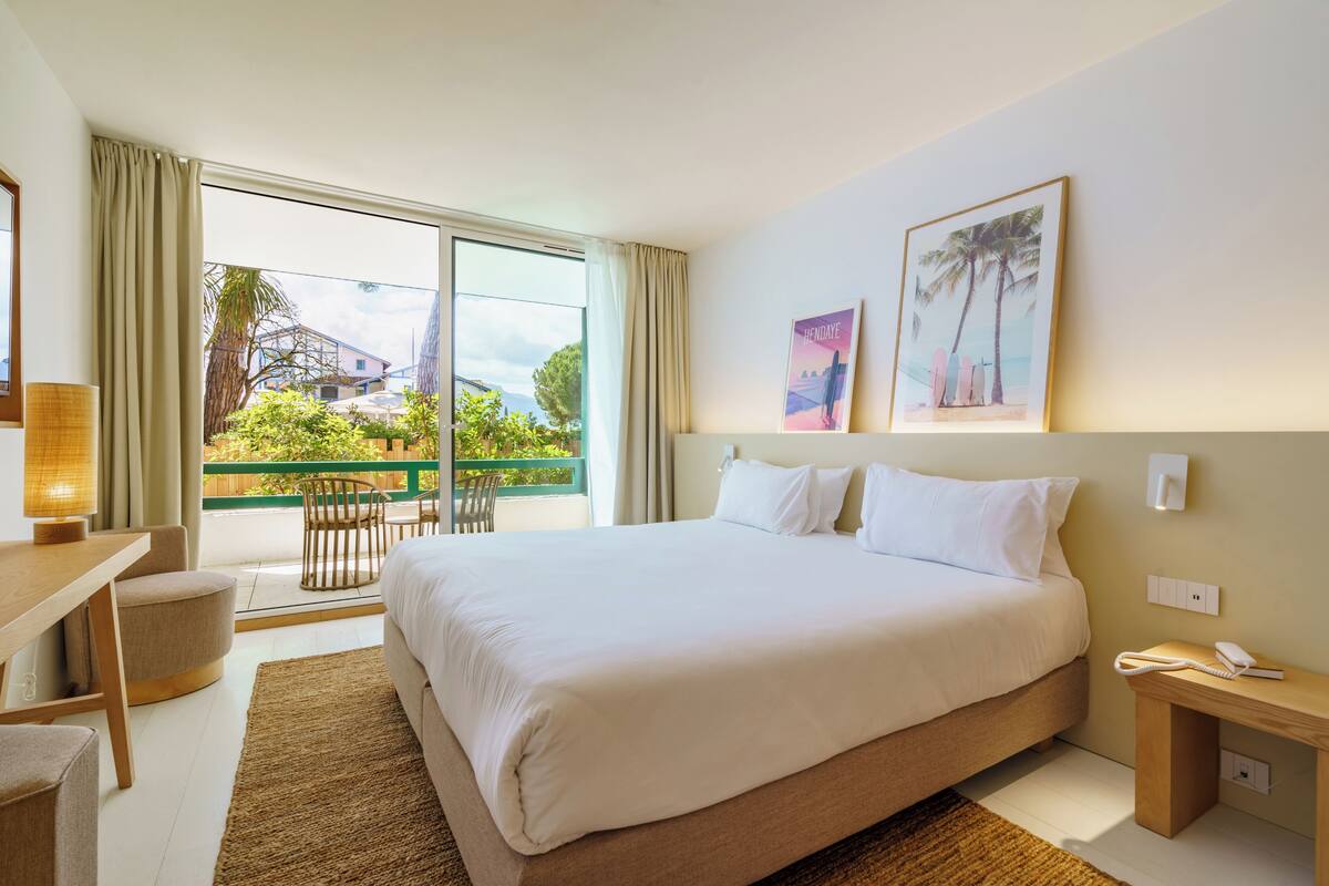 Habitación de hotel con una cama grande, un escritorio, una silla y un balcón con vistas a zonas verdes y edificios.