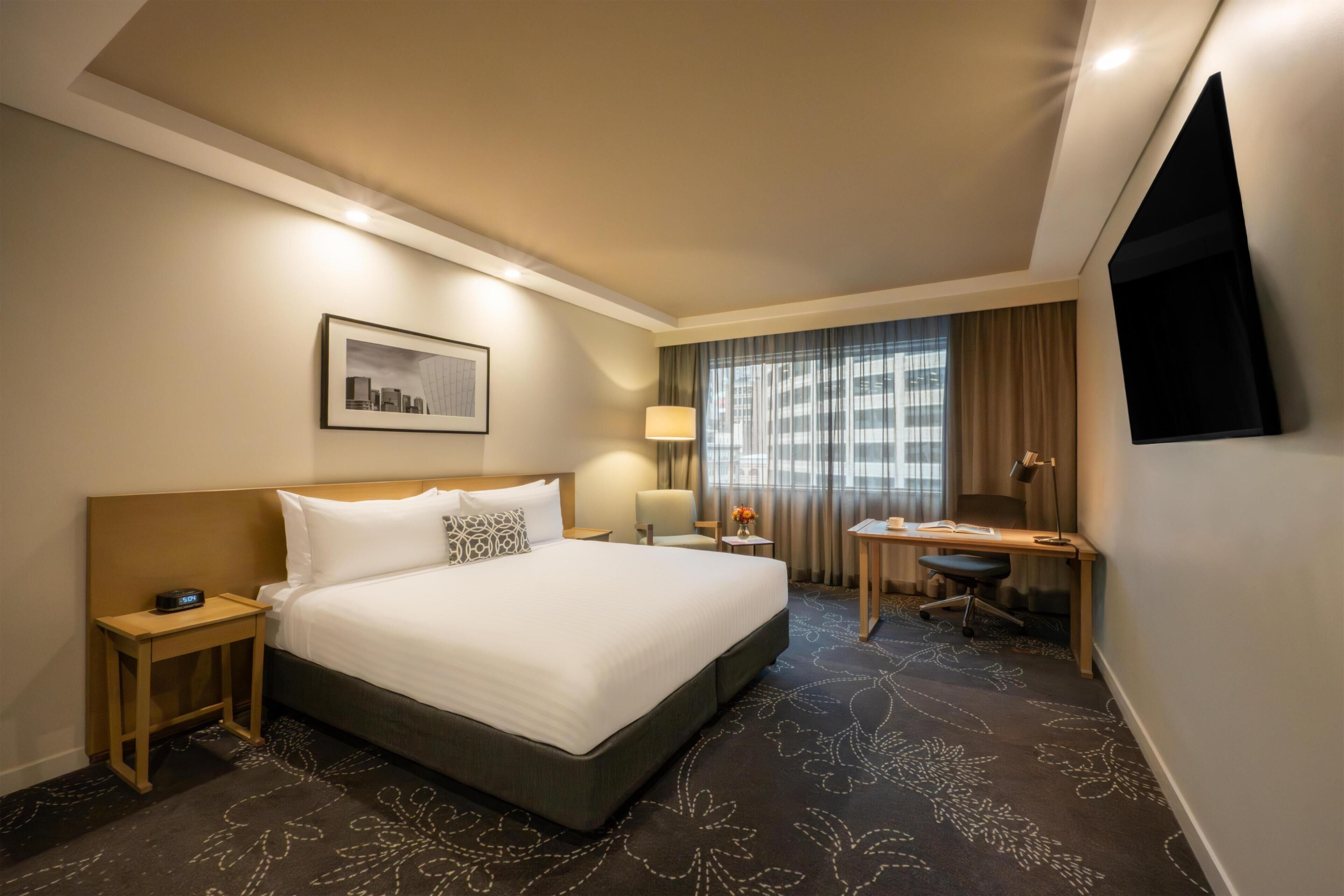 A hotel room with a large bed, a desk, a chair, a TV, a nightstand, a lamp, and a framed picture on the wall.