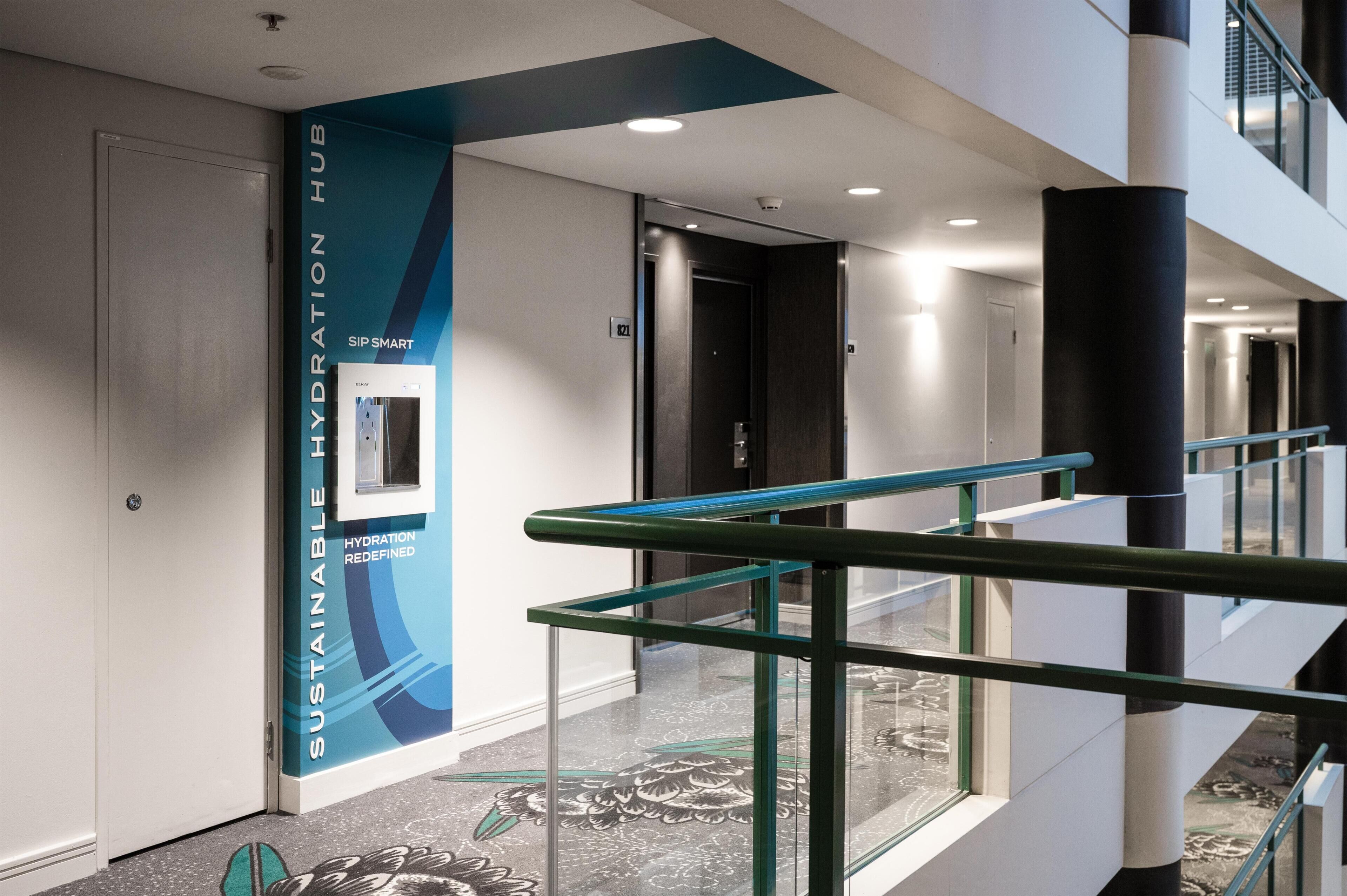 Een moderne hotelgang met een glazen balustrade, een waterdispenser en een bordje voor de 'Sustainable Hydration Hub'.