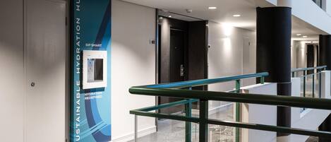 Een moderne hotelgang met een glazen balustrade, een waterdispenser en een bordje voor de 'Sustainable Hydration Hub'.