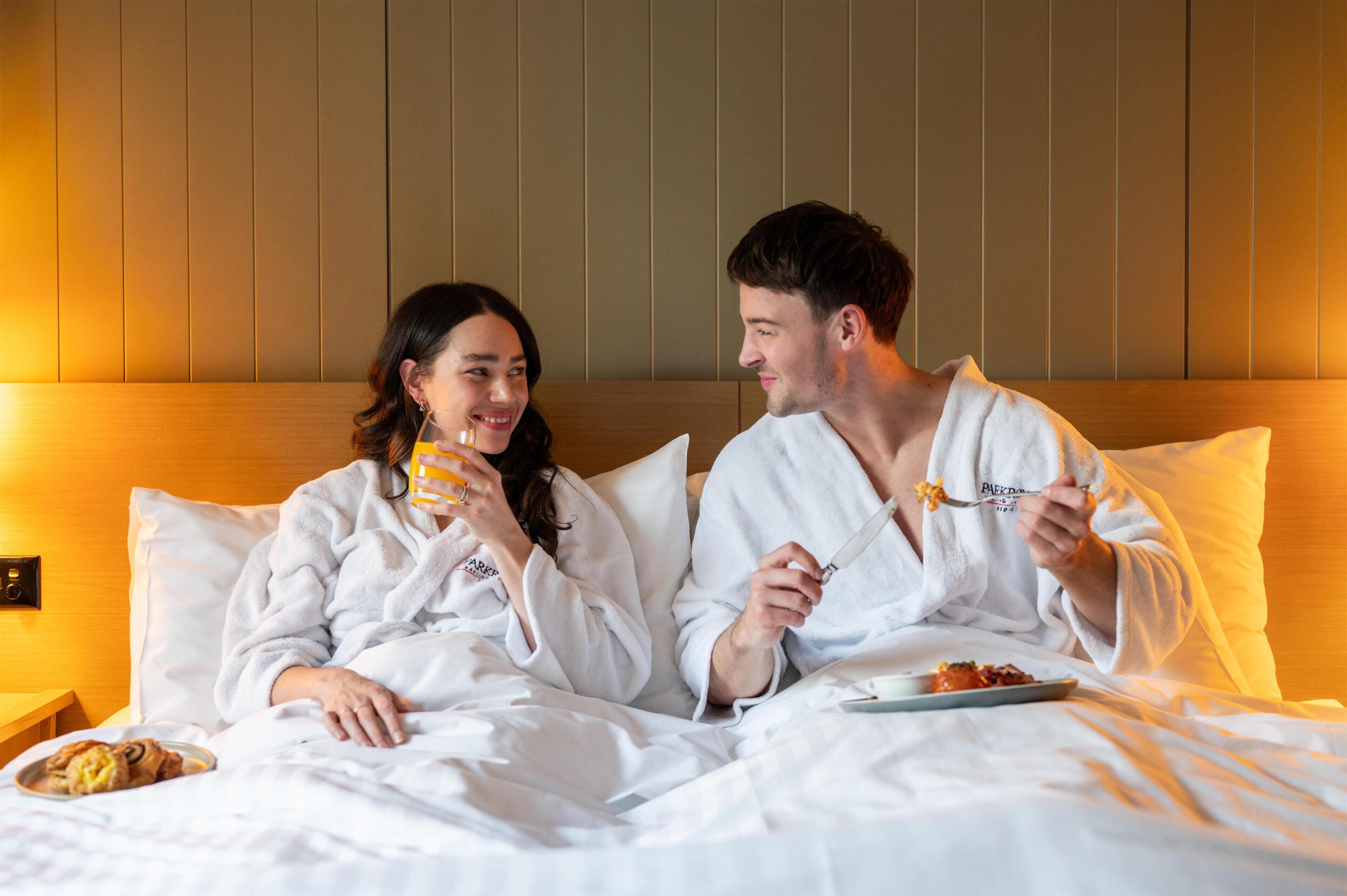 A couple in a hotel room with a bed, pillows, and a bedside table with a plate of food.