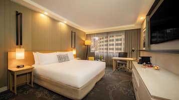 A hotel room with a large bed, bedside tables with lamps, a desk, a television, and a small kitchenette area.