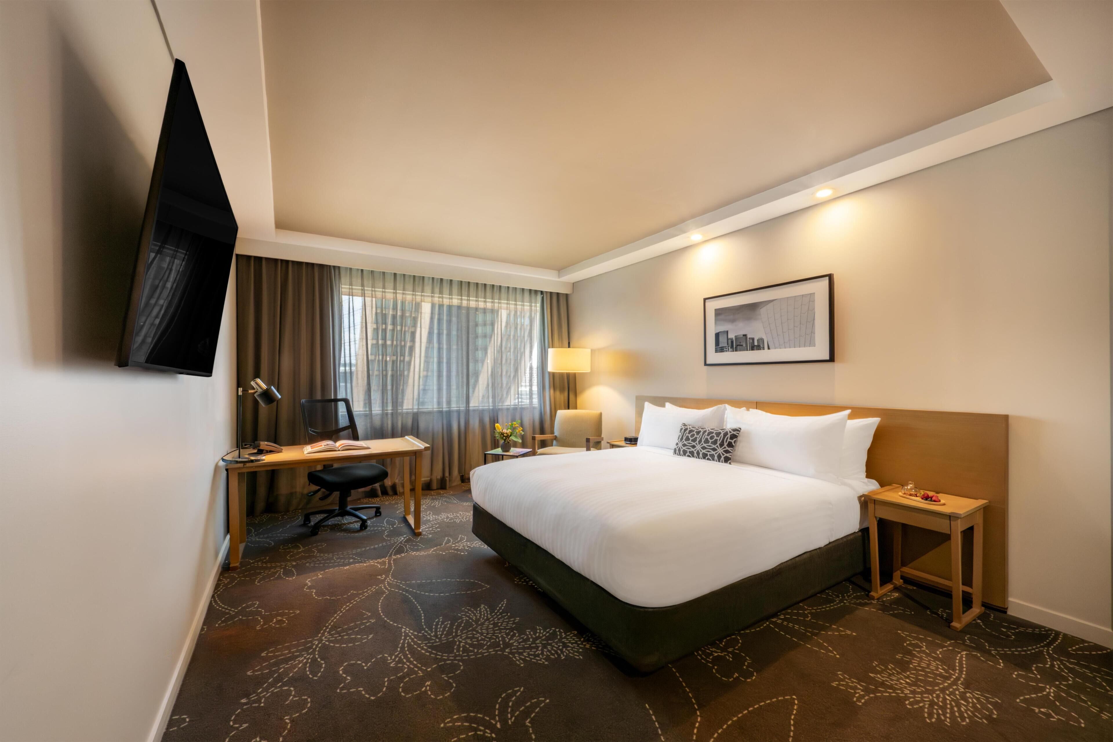 Quarto de hotel com uma cama grande, uma escrivaninha com cadeira, televisão montada na parede e uma janela com cortinas.
