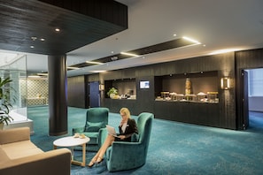 Un moderno vestíbulo de hotel con alfombra color turquesa, una zona de estar con una mesa redonda y un pilar grande.
