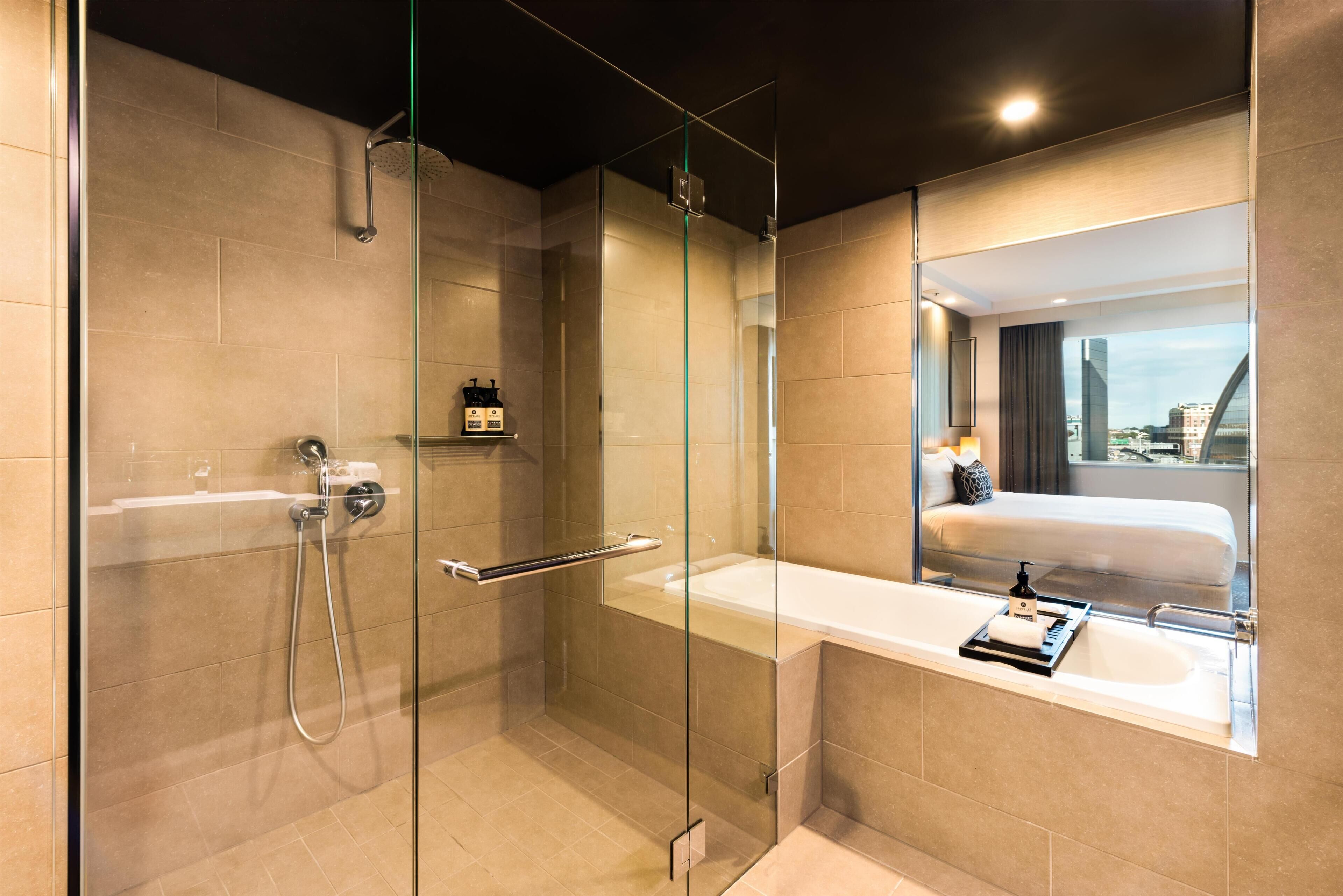 A modern bathroom with a glass-enclosed shower, a large mirror, and a view of a bed and cityscape through the window.