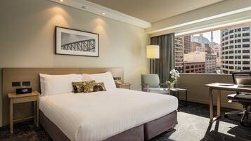 A hotel room with a large bed, a desk, a chair, a lamp, a picture on the wall, and a view of the city.