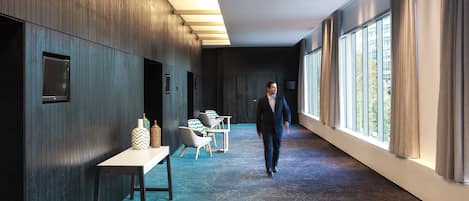 A modern hotel corridor with a long table and chairs, large windows, and a patterned carpet.