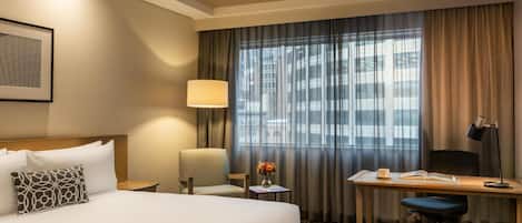 Habitación de hotel con cama, escritorio, silla, lámpara y vistas a la ciudad desde la ventana.