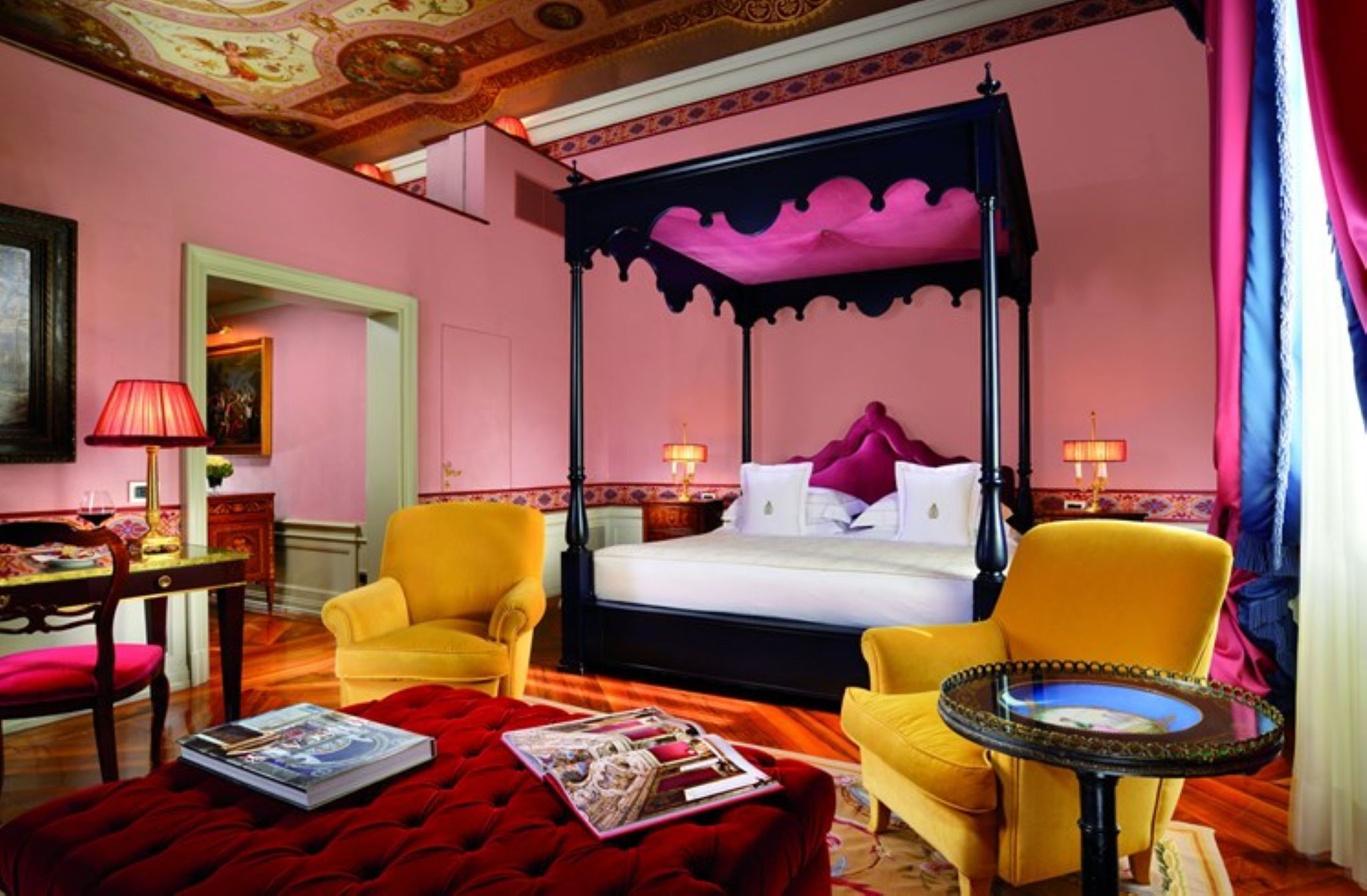 A four-poster bed with a canopy, two yellow armchairs, a red ottoman, and a small round table with a glass top.