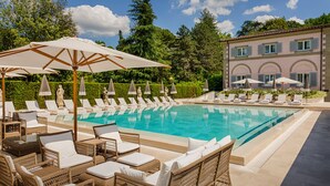 A poolside area with lounge chairs and umbrellas, a clear pool, and a building with arched windows in the background.