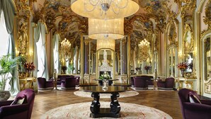 A grand room with ornate gold detailing, multiple chandeliers, and a central table with a vase of flowers.