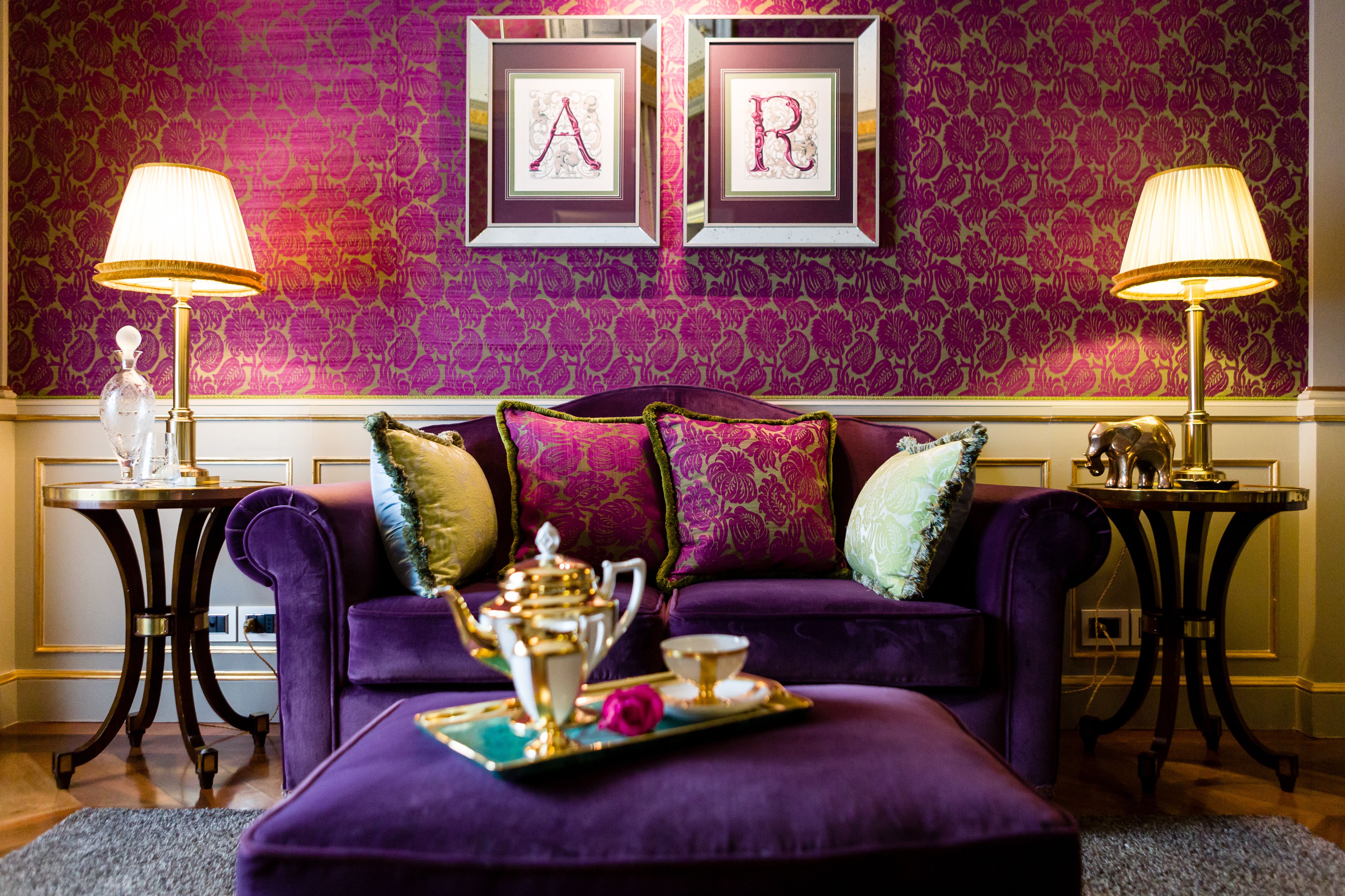 Um sofá roxo com almofadas decorativas, um tabuleiro com bule e xícaras, e duas molduras com as letras 'A' e 'R' na parede.