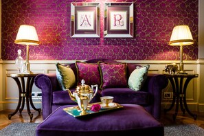A purple sofa with decorative pillows, a tray with a teapot and cups, and two framed letters 'A' and 'R' on the wall.