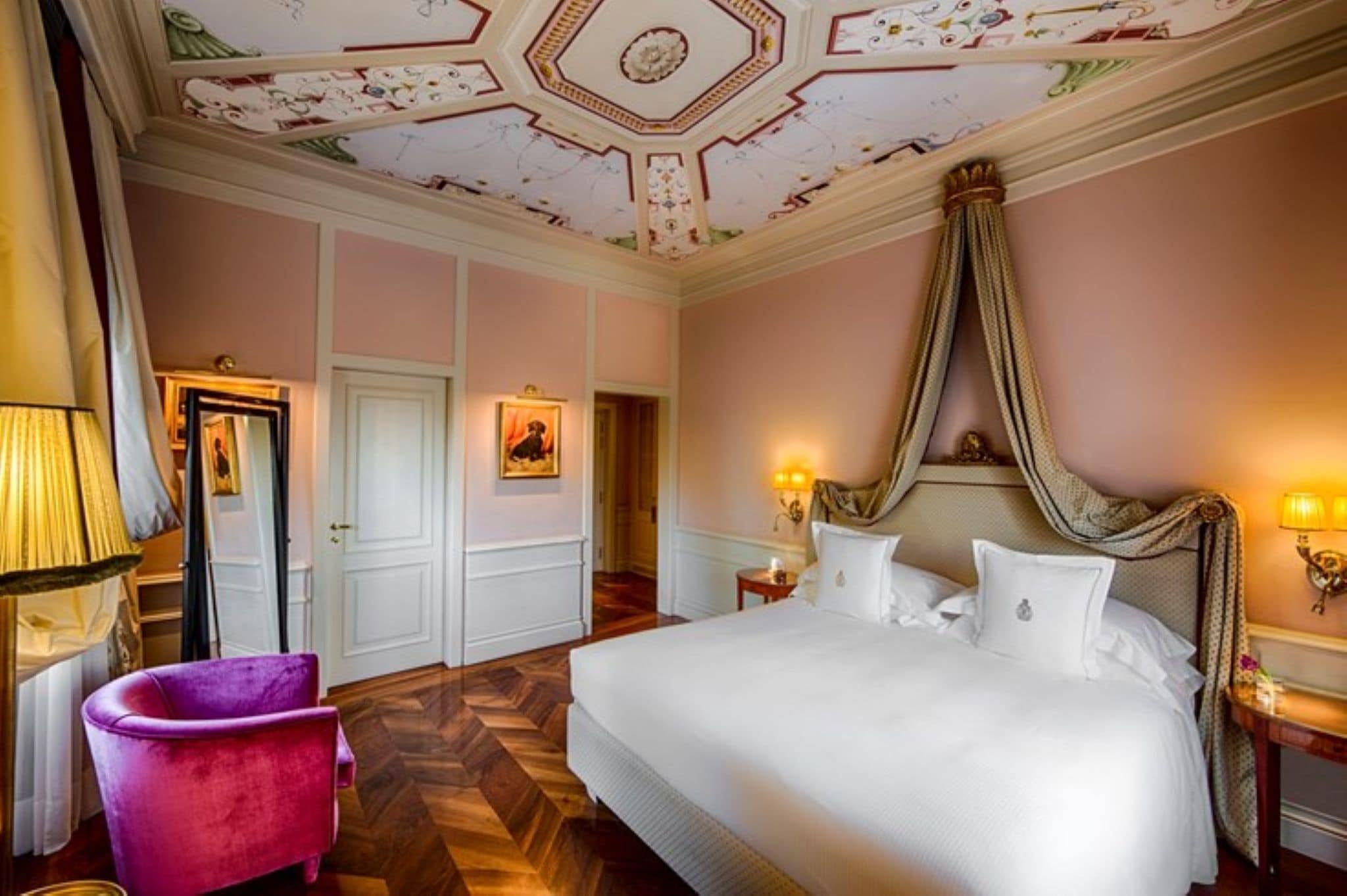 A hotel room with a large bed, a vanity with a mirror, a pink armchair, and a decorative ceiling.