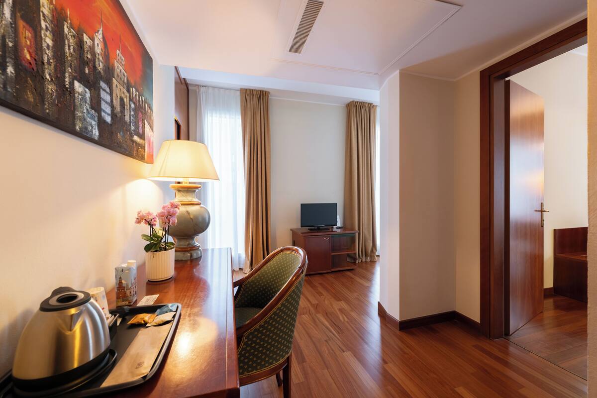 Una camera d'albergo con pavimento in legno, una scrivania con lampada, una sedia, una televisione e un quadro appeso al muro.