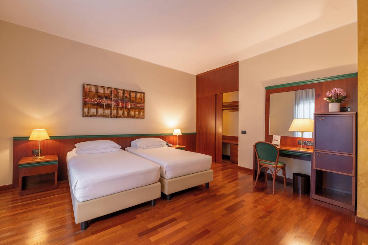 Camera d'albergo con due letti, pavimento in legno, una scrivania con una sedia, uno specchio e un quadro appeso al muro.