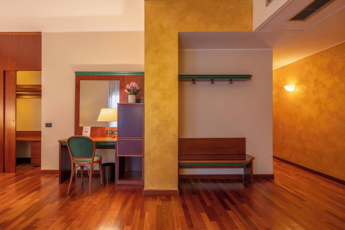 Camera d'albergo con scrivania in legno, una sedia, una panca, un cestino e un lampadario a parete.