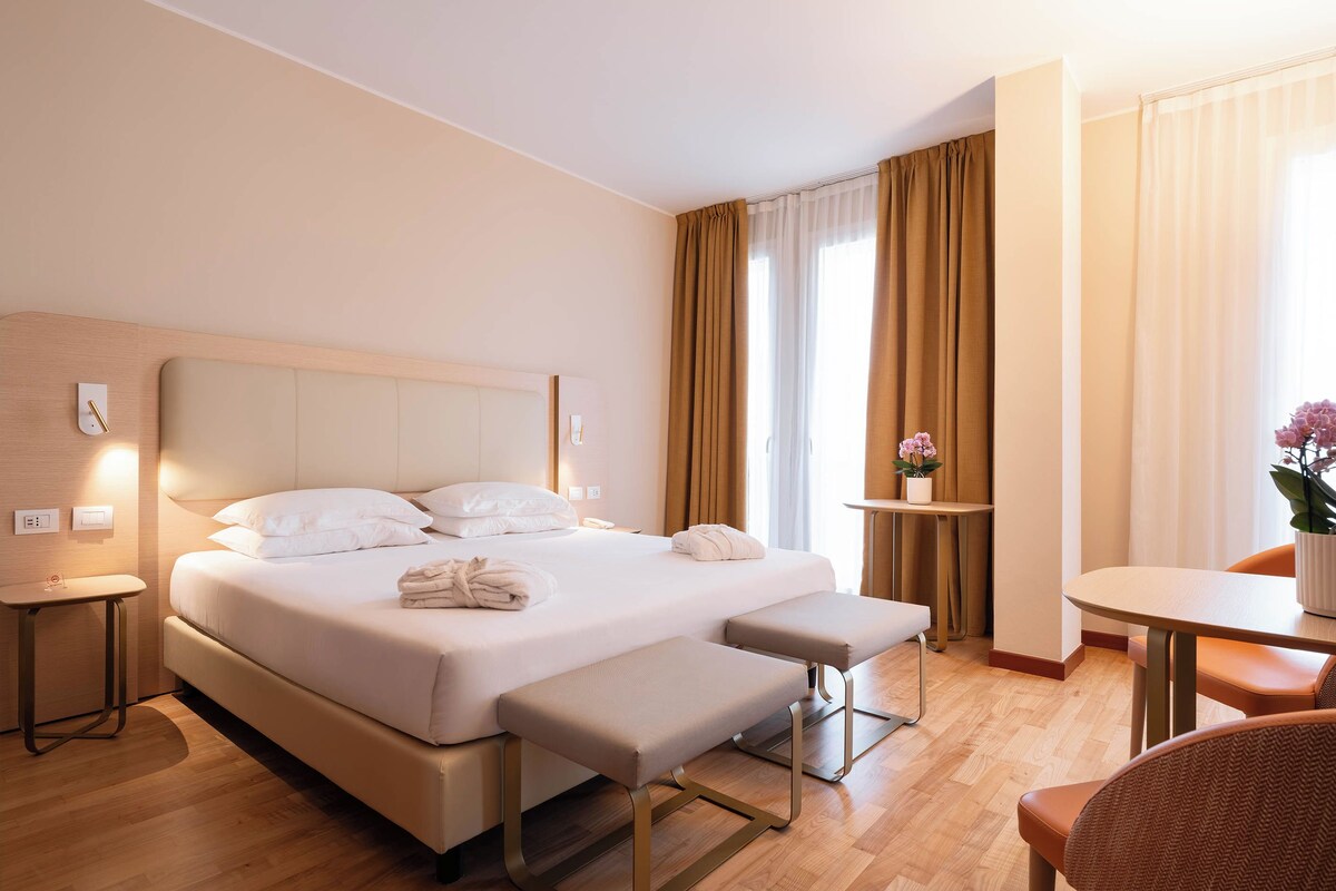 A hotel room with a large bed, bedside tables, a small table with a vase, and a chair.