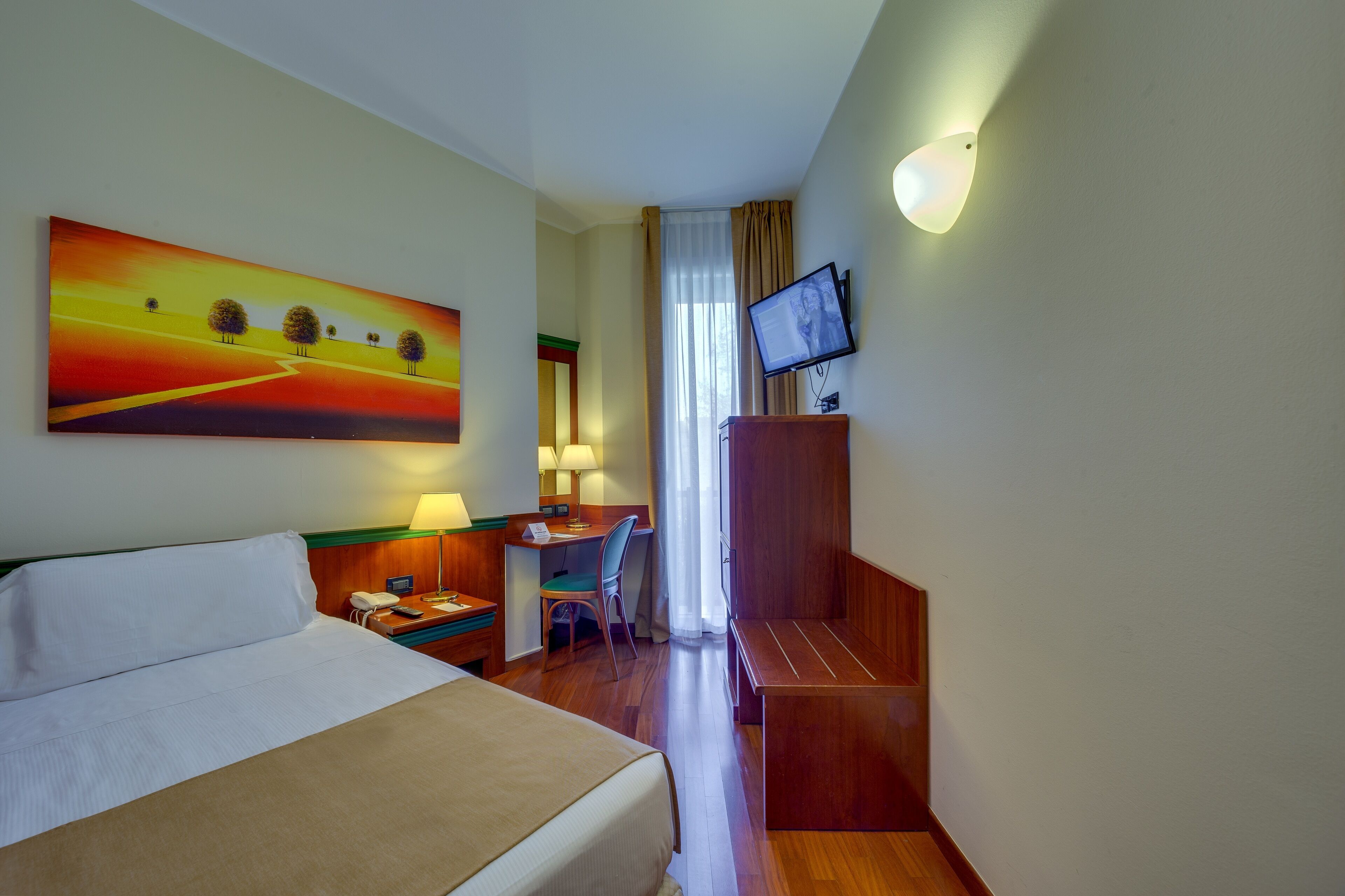 A hotel room with a bed, desk, chair, TV, and a painting on the wall.