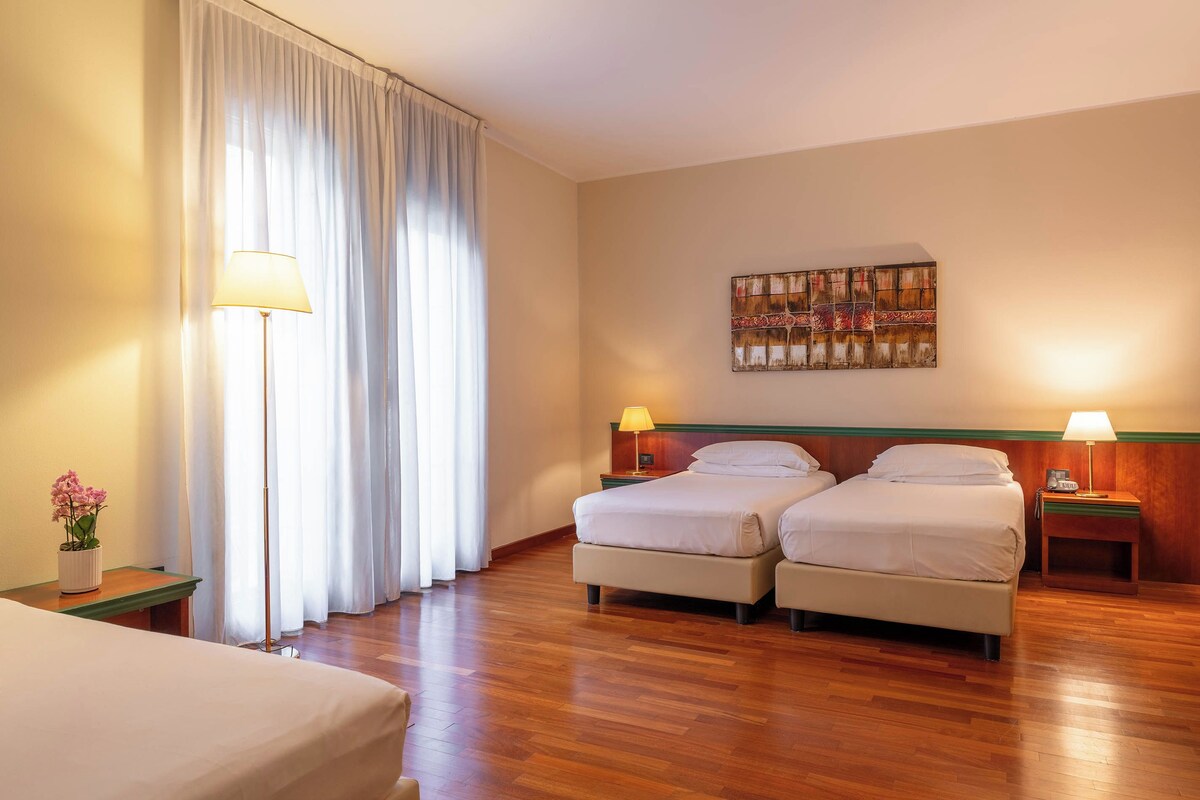 A hotel room with two beds, a large window with curtains, a painting on the wall, and a bedside table with a lamp.