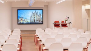 A conference room with rows of white chairs, a large screen displaying a cityscape, and a podium with a vase of flowers. - Hotel Raffaello (Milan)