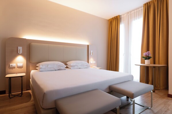 Habitación de hotel moderna con una cama grande, mesitas de noche, una mesita pequeña con una planta y un ventanal con cortinas.
