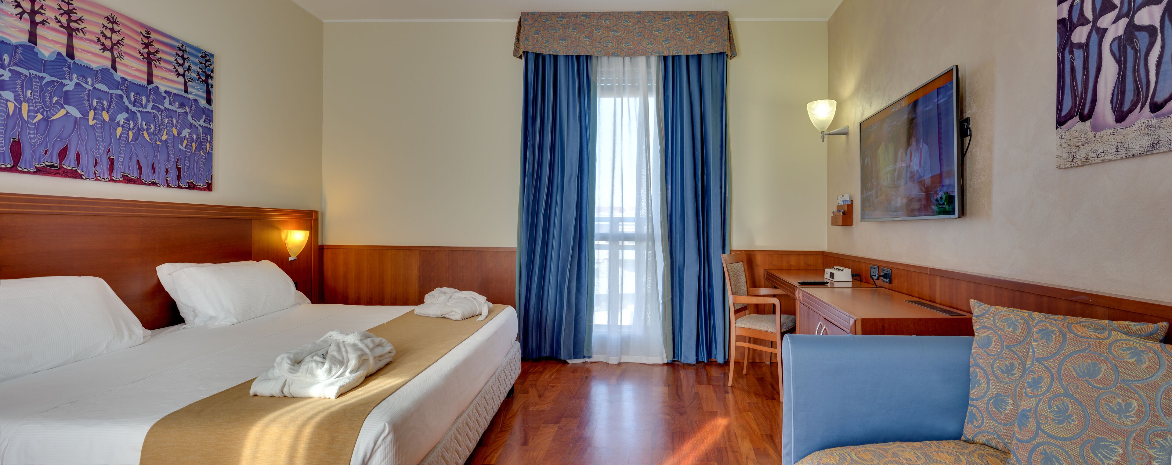 A hotel room with a bed, a desk, a chair, and a large window with blue curtains.