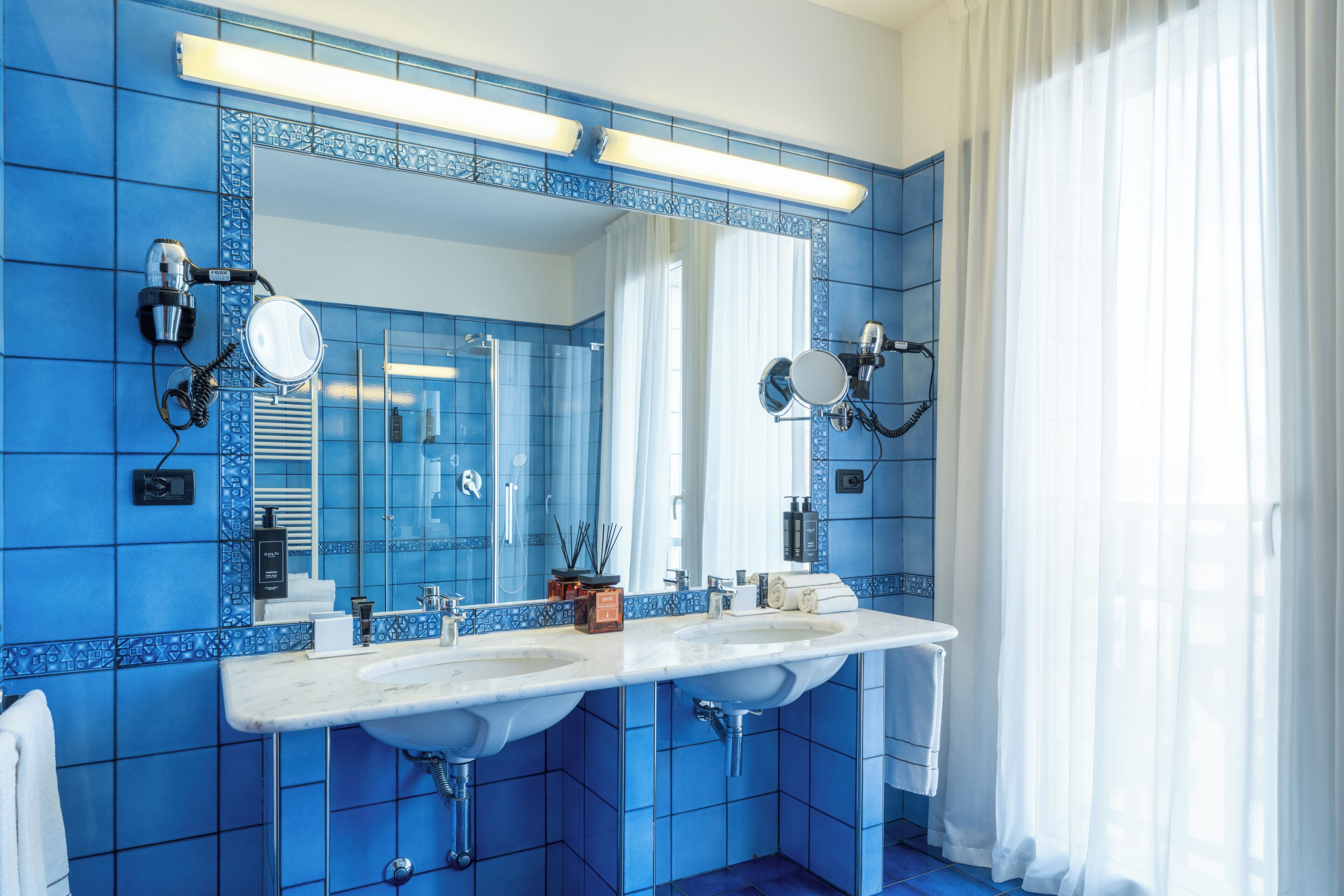 Une salle de bain avec des carreaux bleus, deux lavabos, un grand miroir et une fenêtre avec des rideaux blancs.