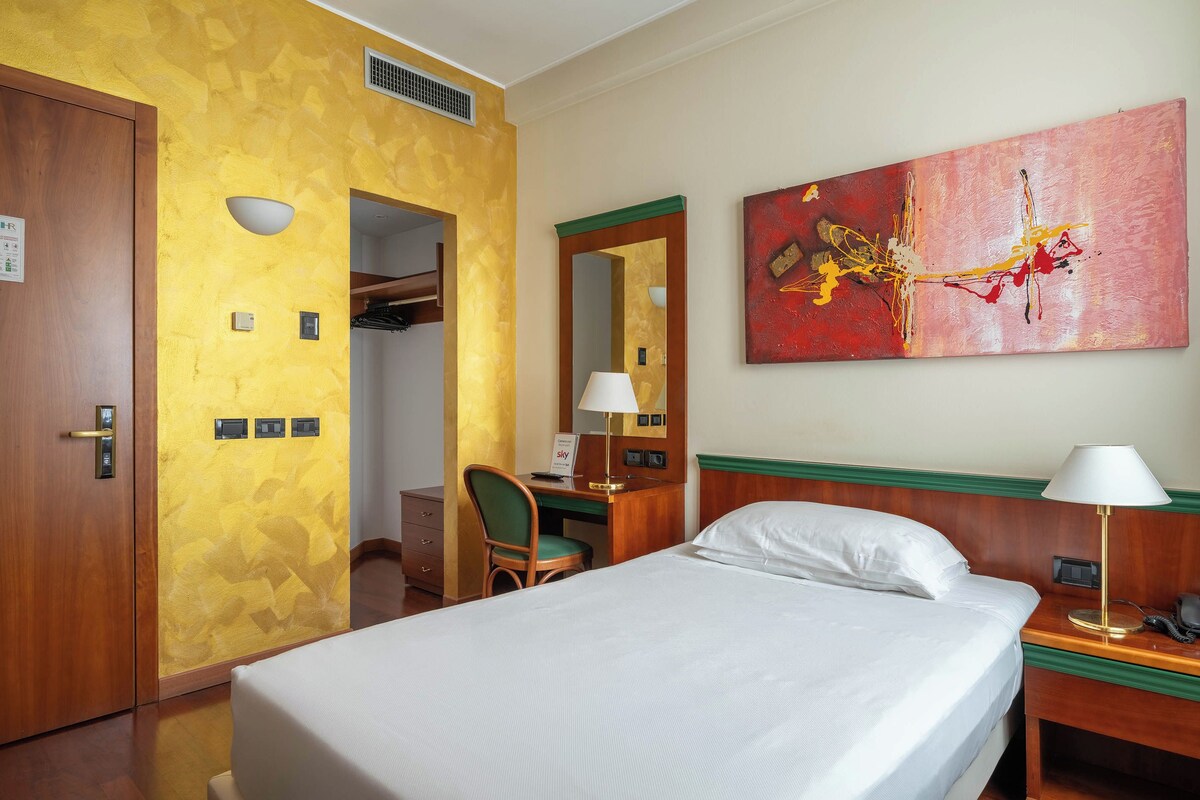 A hotel room with a bed, a desk with a chair, a desk lamp, a telephone, and a painting on the wall.