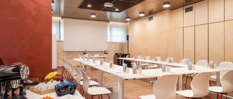 A conference room with white tables and chairs, a projector screen, and a buffet table with pastries and drinks.