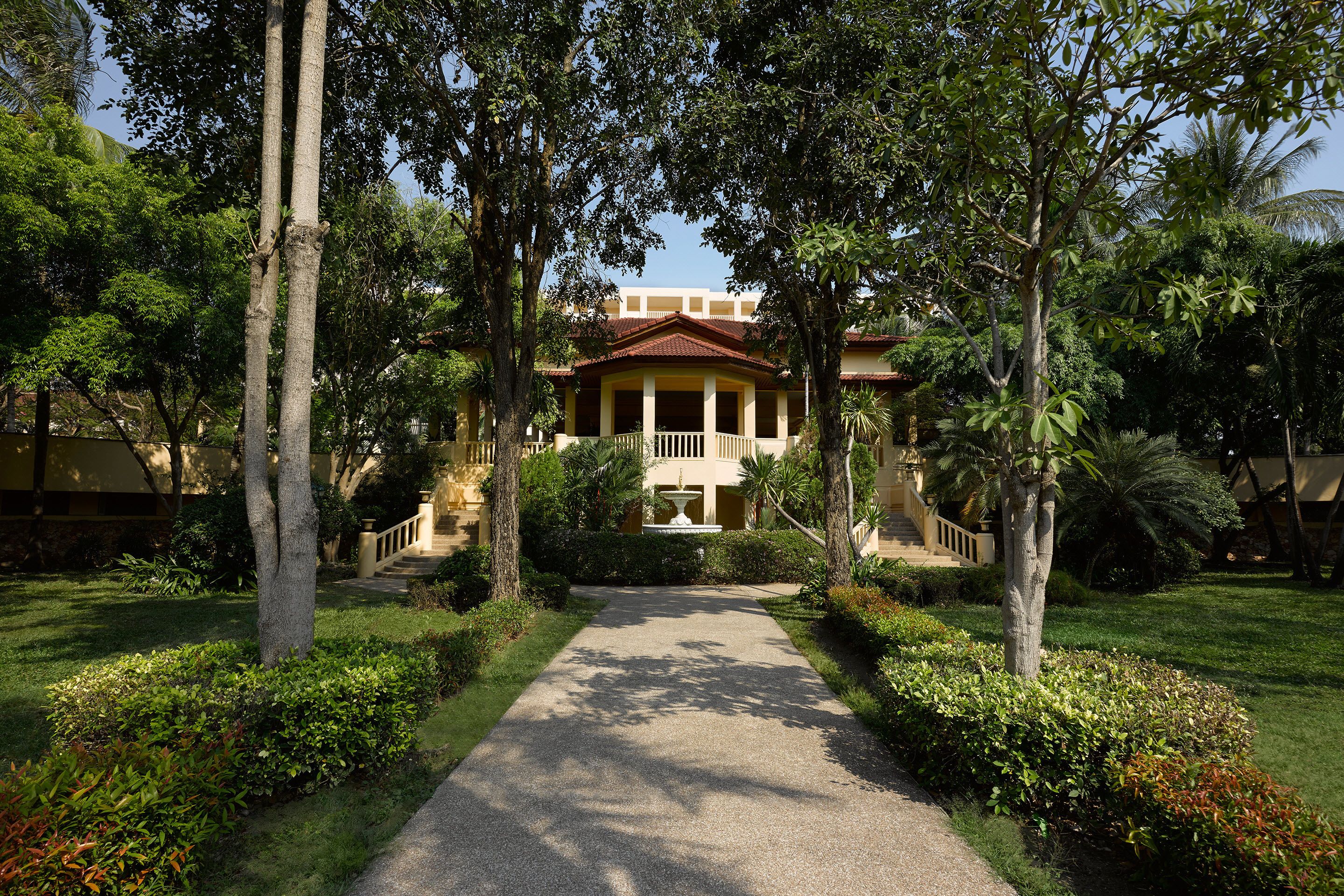 A pathway leading to a building with a central fountain, surrounded by well-maintained greenery and trees.