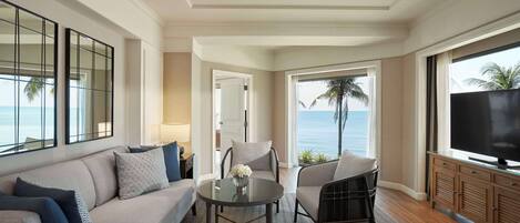 A modern living room with a sofa, chairs, a coffee table, and a TV. There is a view of the ocean through large windows.