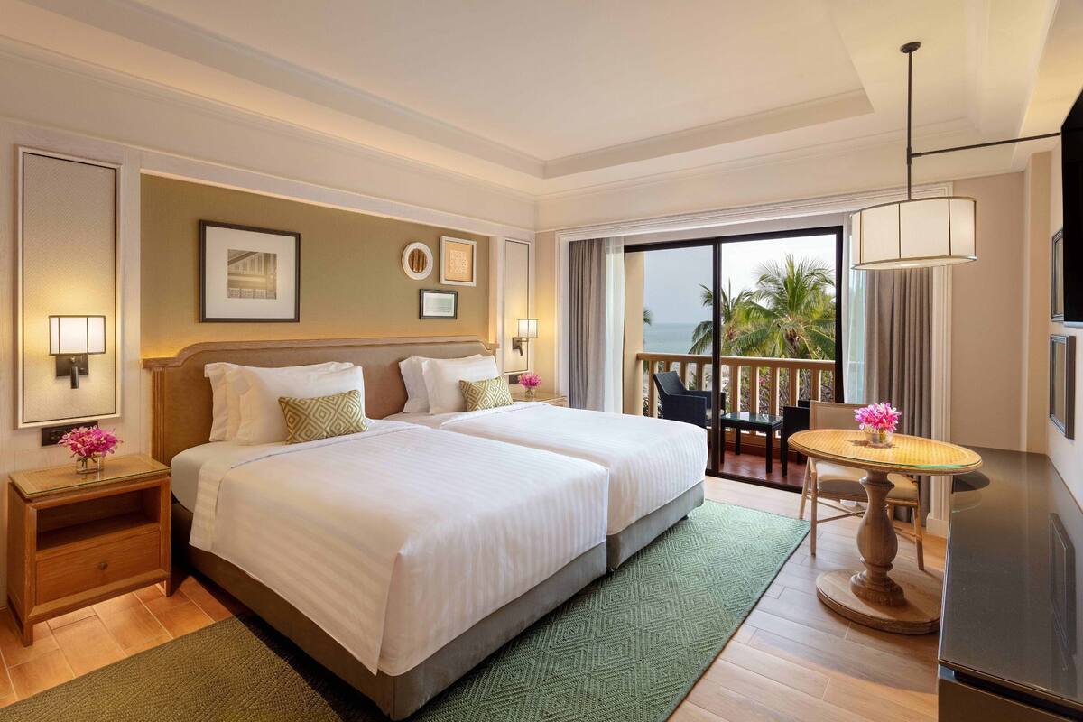 Habitación de hotel con dos camas, una mesa de comedor, un balcón con vista a palmeras y un ventanal grande.
