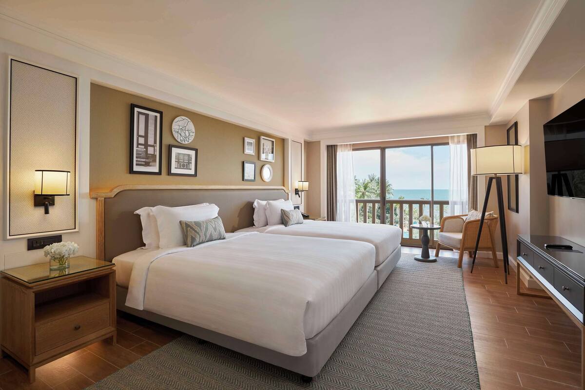 Habitación de hotel con dos camas, televisión, escritorio y balcón con vistas.