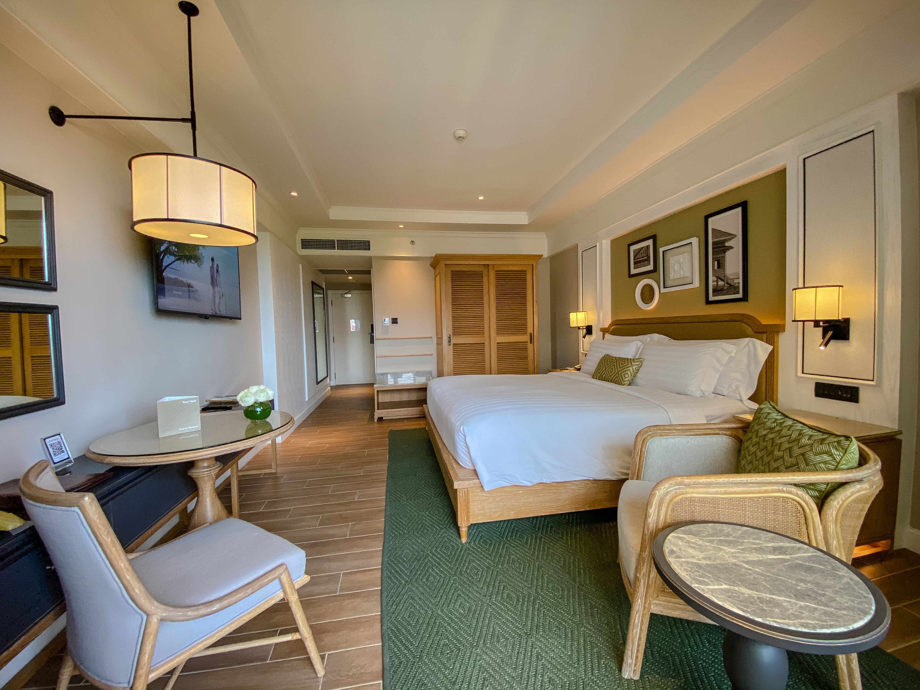 Habitación de hotel con cama, escritorio con silla, pequeña mesa con un florero y alfombra verde.