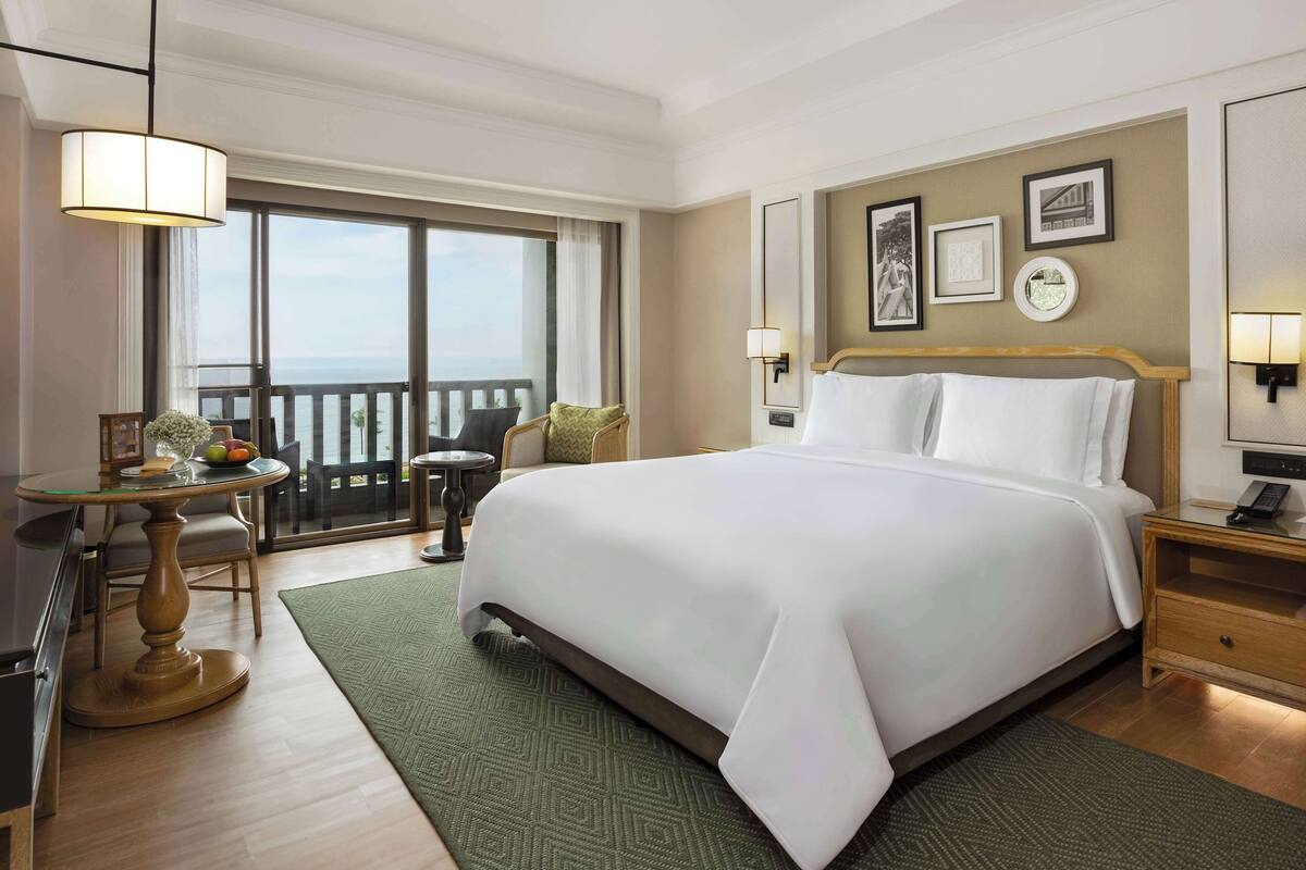 Habitación de hotel con una cama grande, zona de estar con mesa, vista al océano y balcón.