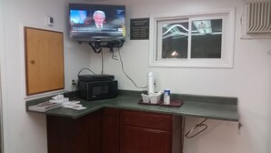 A small kitchenette with a microwave, a coffee maker, and a TV displaying a news broadcast. There is a window with blinds and a sign indicating complimentary breakfast hours. - Rapids Inn and Suites (Wisconsin Rapids)