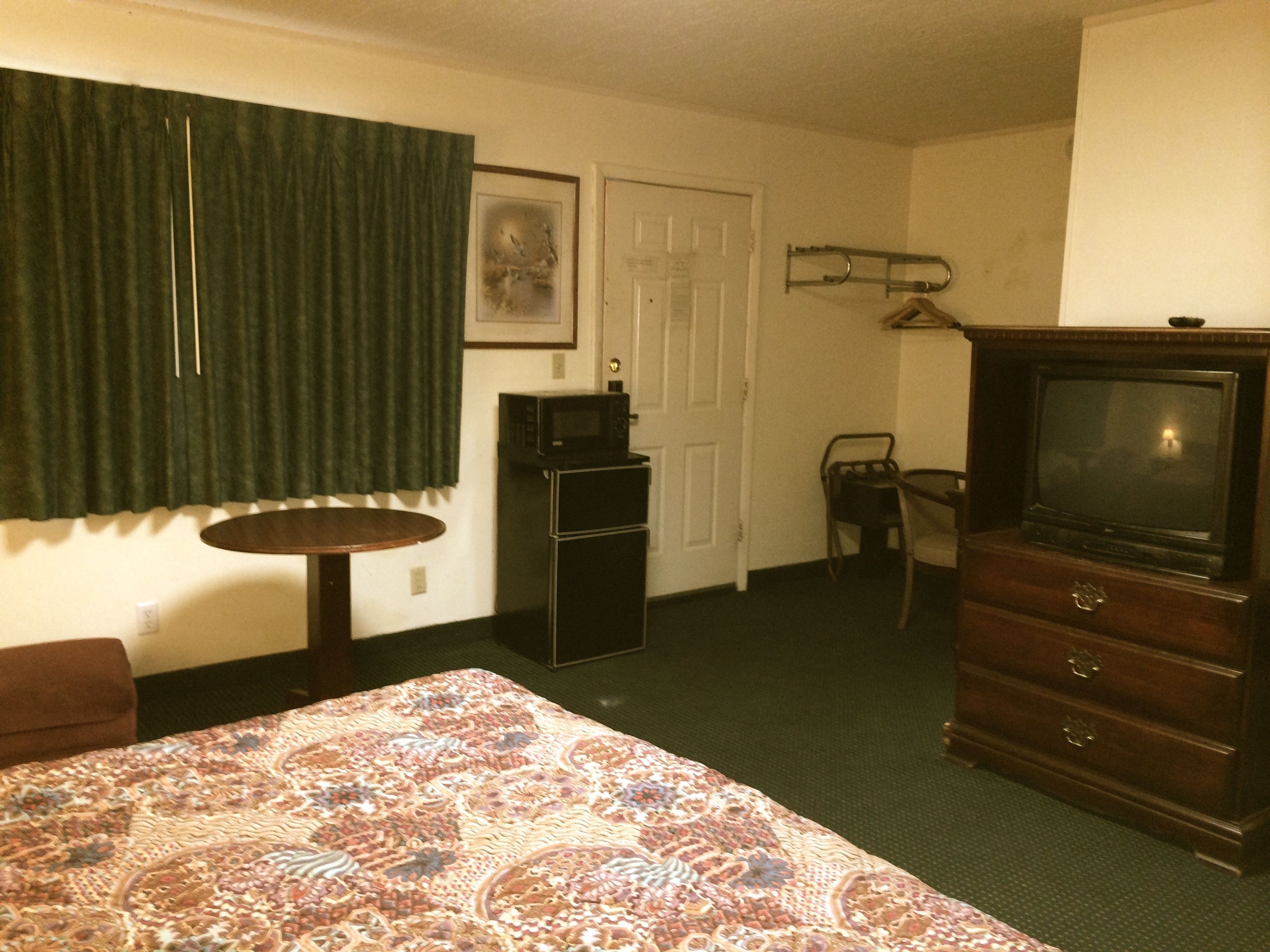 A hotel room with a bed, a television, a small table, a chair, a door, and a coat rack.